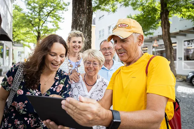 Ältere und jüngere Menschen schauen lächelnd auf ein Tablet auf einer Straße