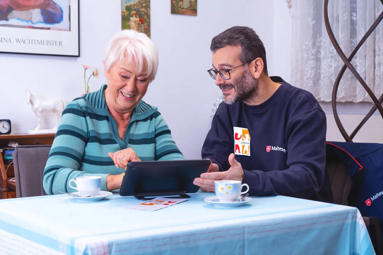 Eine ältere Frau tippt auf einem Tablet. Ein jüngerer Mann unterstützt sie. Sie sitzen lächelnd am Tisch.