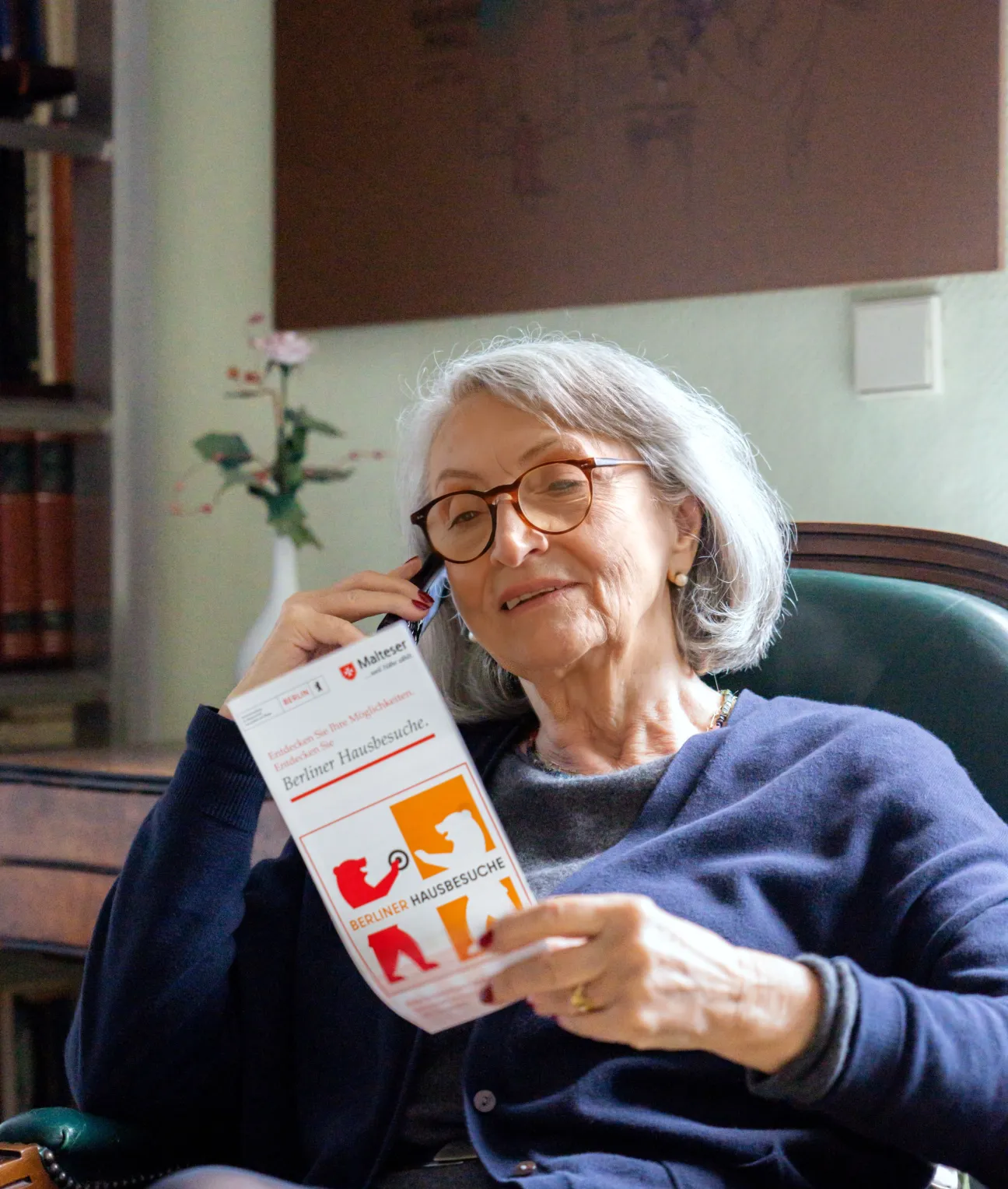 Eine ältere Frau sitzt und telefoniert. Sie betrachtet dabei den Flyer in ihrer Hand.