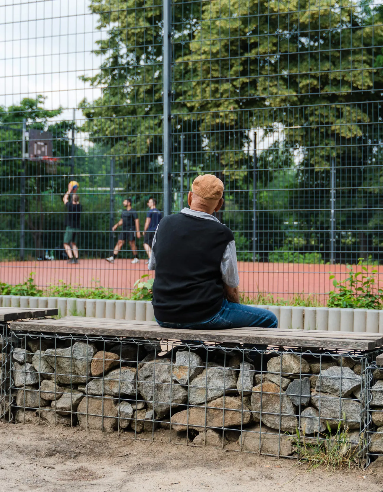 Älterer Mann sitzend von hinten, einem belebtem Basketbalplatz zuwandt.