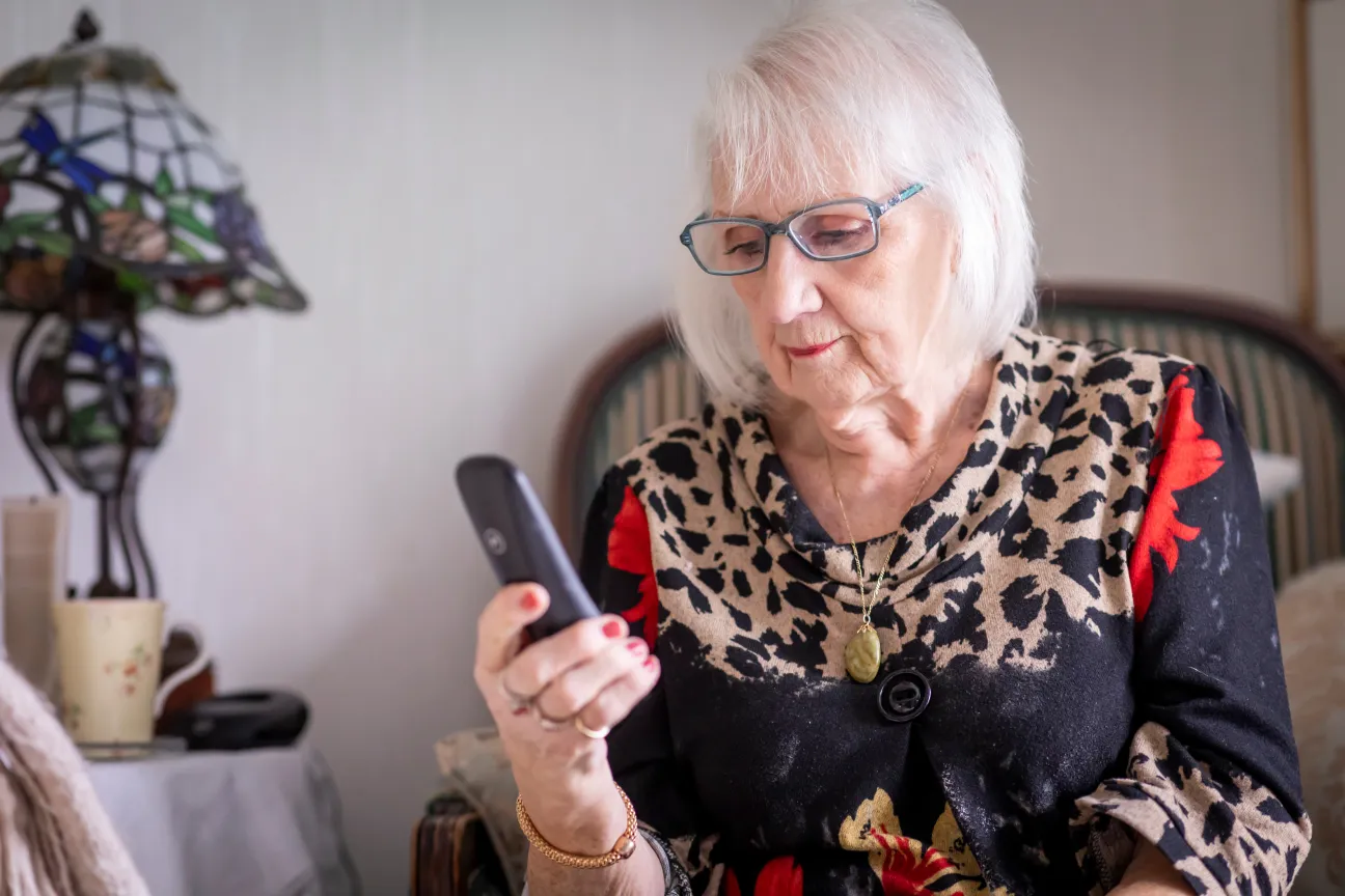 Eine ältere Frau sitzt in einem Wohnzimmer und tippt in ihr Telefon.