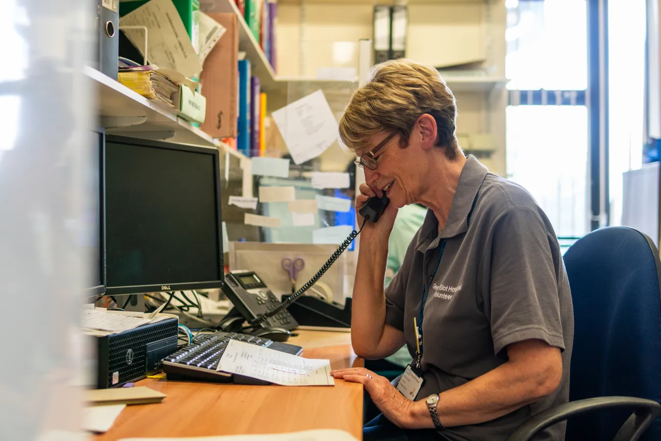 Eine Frau mittleren Alters sitzt in einer Telefonzentrale und spricht freundlich am Telefon.