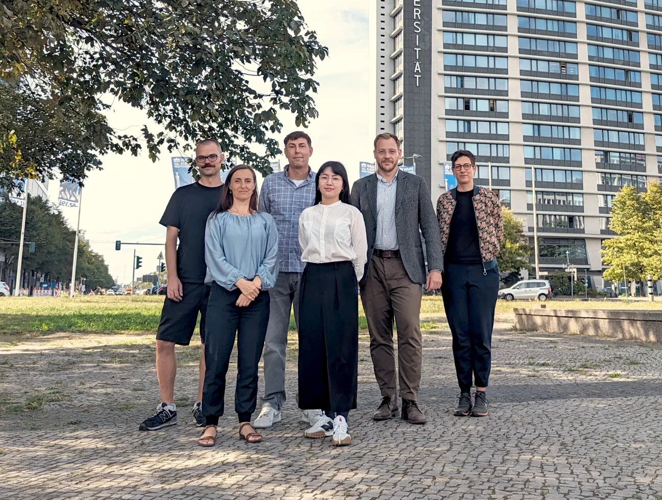 3 Frauen und 3 Männer stehen zusammen für ein Gruppenbild auf Pflastersteinen vor einem Hochhaus.