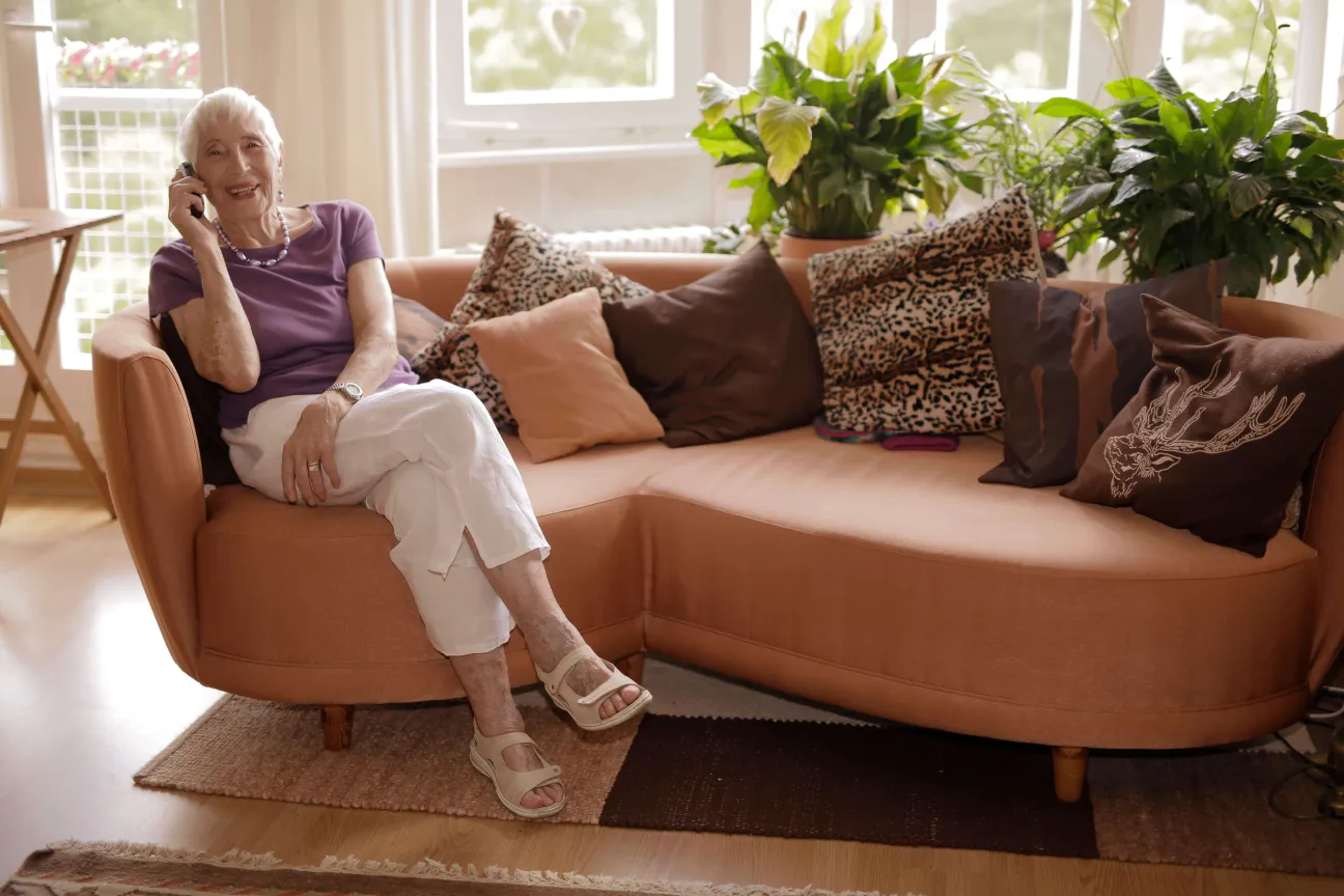 Ältere Frau sitzt auf einem Sofa und telefoniert lächelnd.