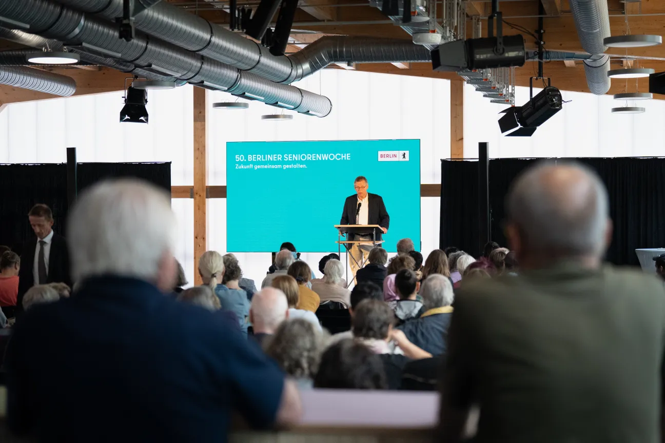 Menschen sitzen in einem Saal und schauen zu einem Mann, der vor einer digitalen Leinwand am Pult steht und spricht.
