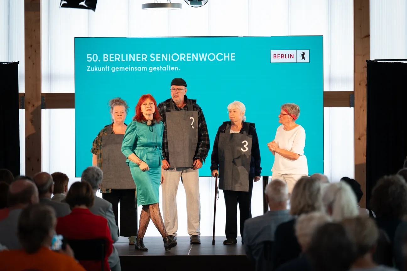 Menschen sitzen auf Stühlen und schauen auf eine Bühne. Dort stehen 5 Personen vor einer digitalen Leinwand.