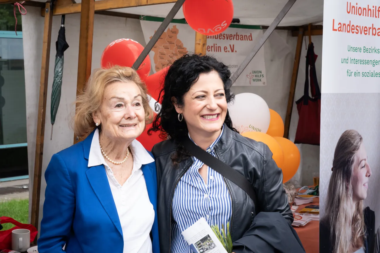 Zwei Frauen stehen dicht nebeneinander vor einem Informationsstand und lächeln.