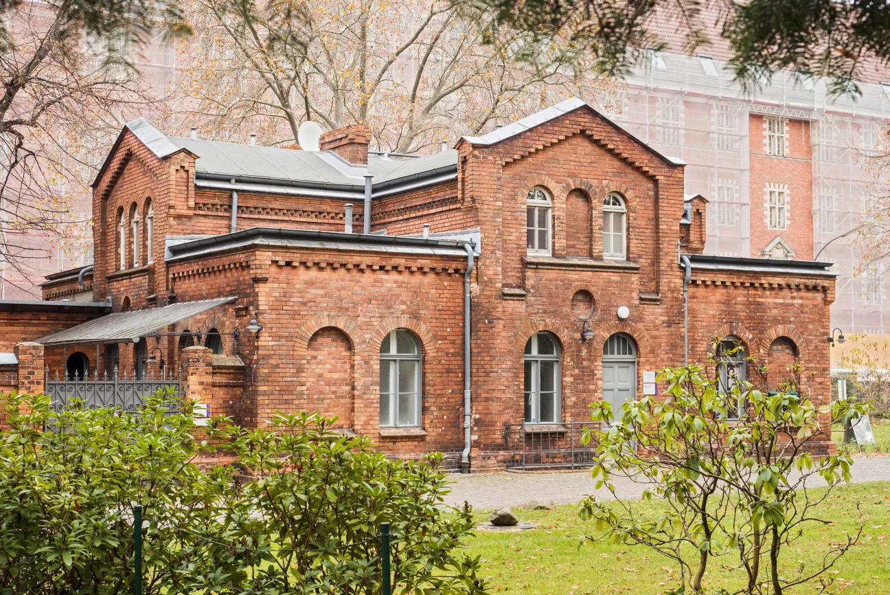 Foto eines zweistöckicgen Backsteinhauses. Davor Wiese und Büsche. Dahinter steht ein höheres Gebäude.