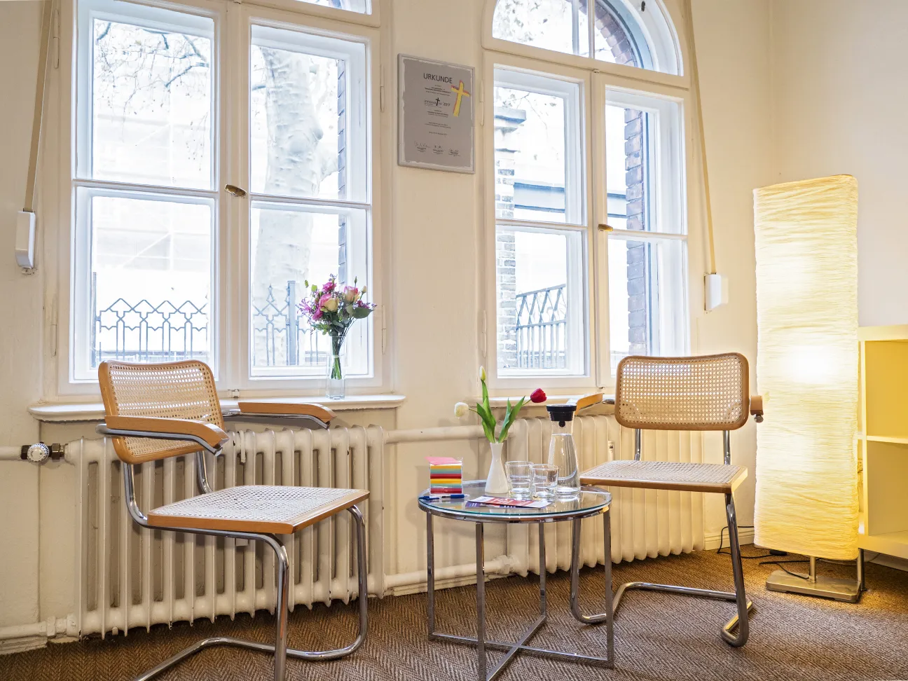 Kleiner Tisch mit zwei Stühlen vor einer Fensterfront. Blumen auf Tisch und Fensterbank. Warmes Licht.