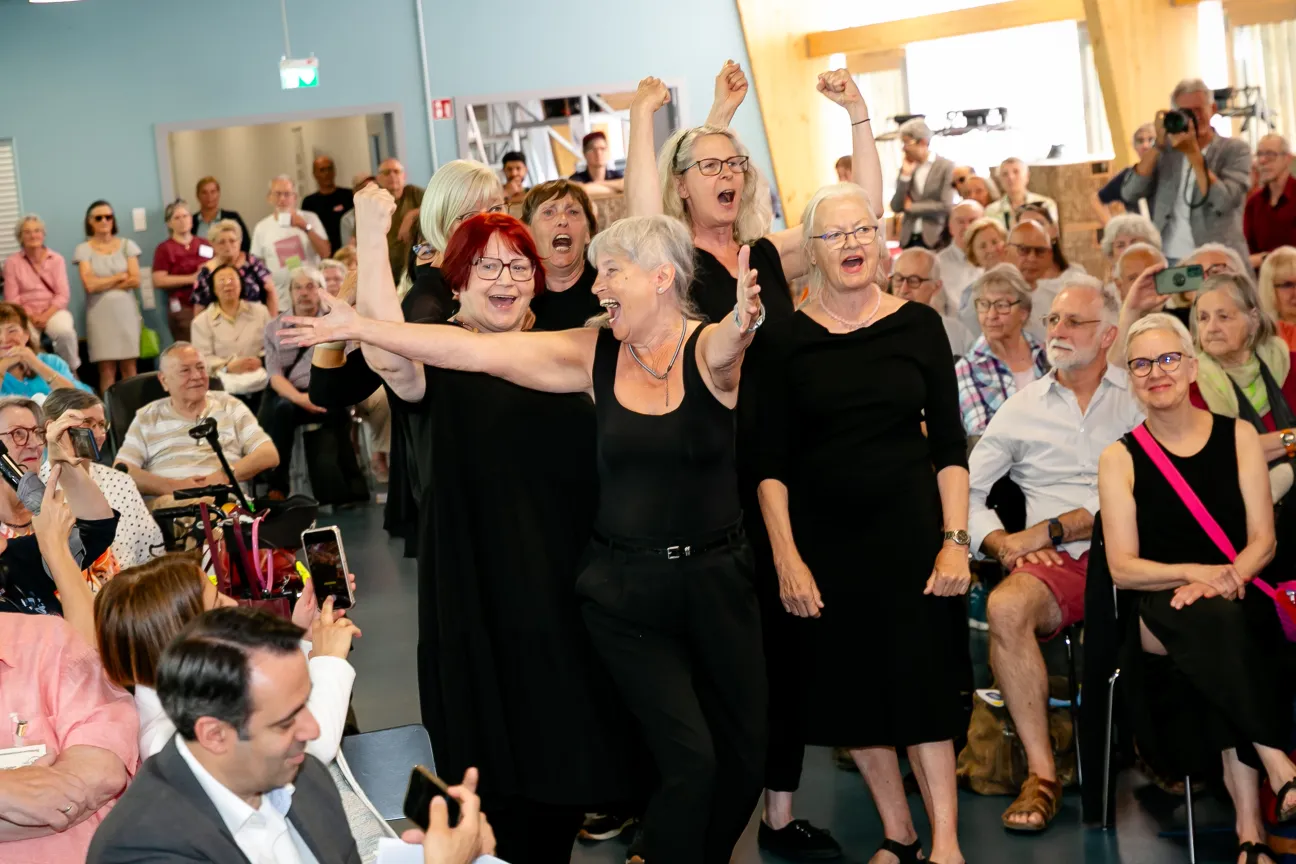 Eine Gruppe älterer Frauen schwarz gekleidet singt fröhlich und hält die Arme hoch. Ringsherum sitzt Publikum und freut sich mit. Einige fotografieren.