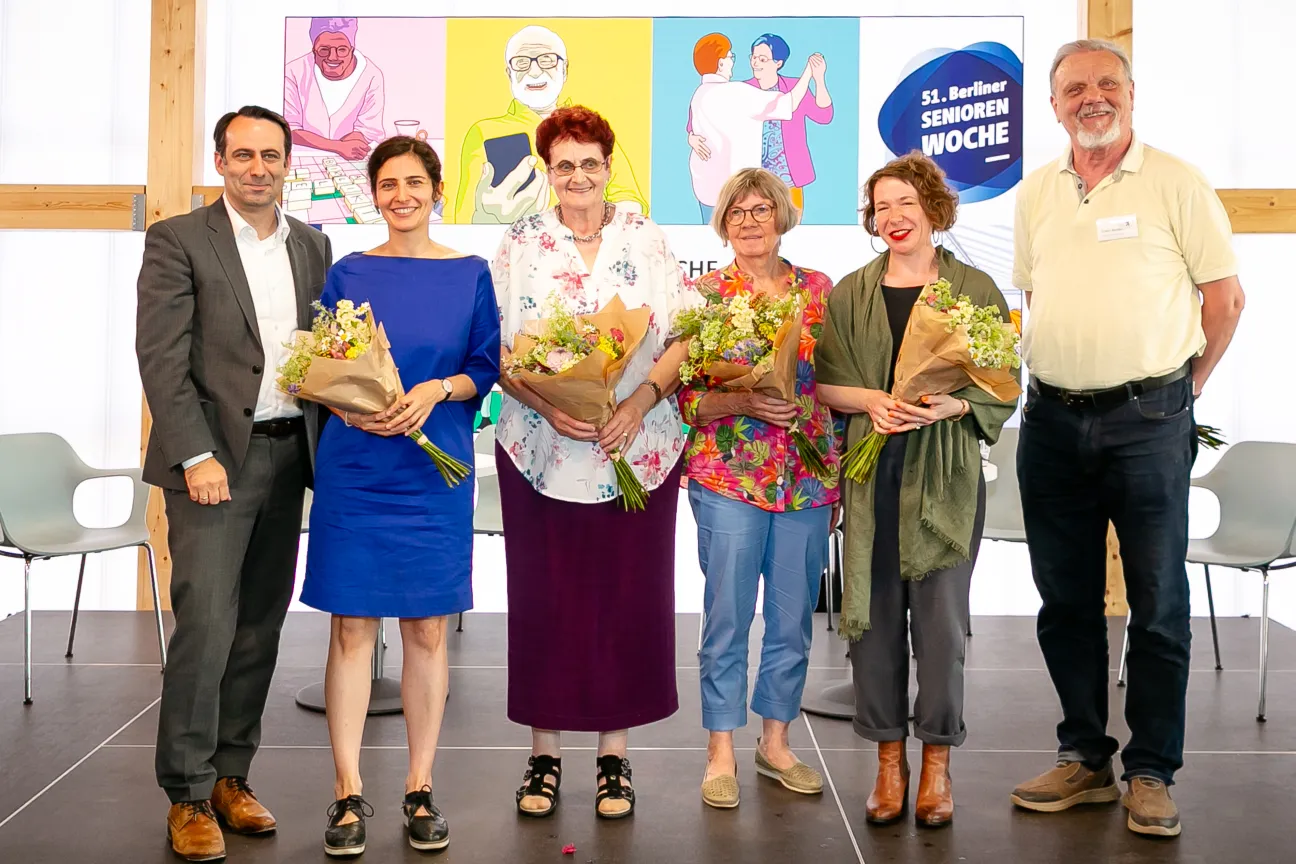 4 Frauen mit Blumensträußen und 2 Männer stehen lächelnd auf einer Bühne. Im Hintergrund bunte Grafiken zur 51. Berliner Seniorenwoche.