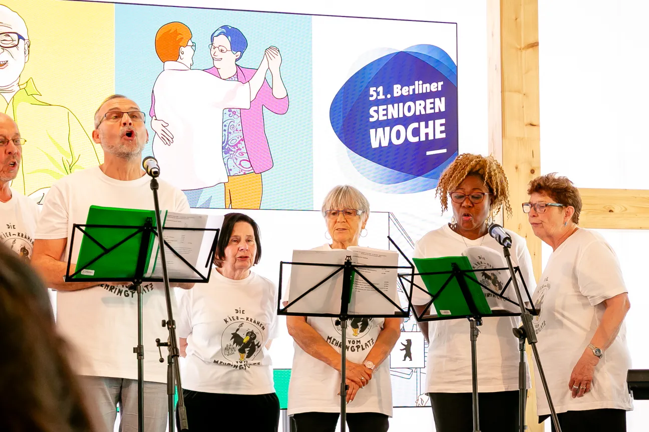 4 Frauen und 2 Männer stehen singend hinter Notenständern. Im Hintergrund eine Grafik tanzender Personen. Zu lesen ist dort: 51. Berliner Seniorenwoche.