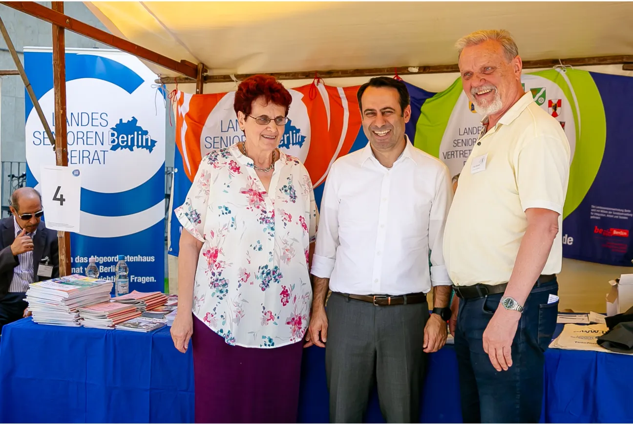 3 lachende Personen stehen vor einem Marktstand des Landesseniorenbeirat Berlin und der Landesseniorenvertretung Berlin.