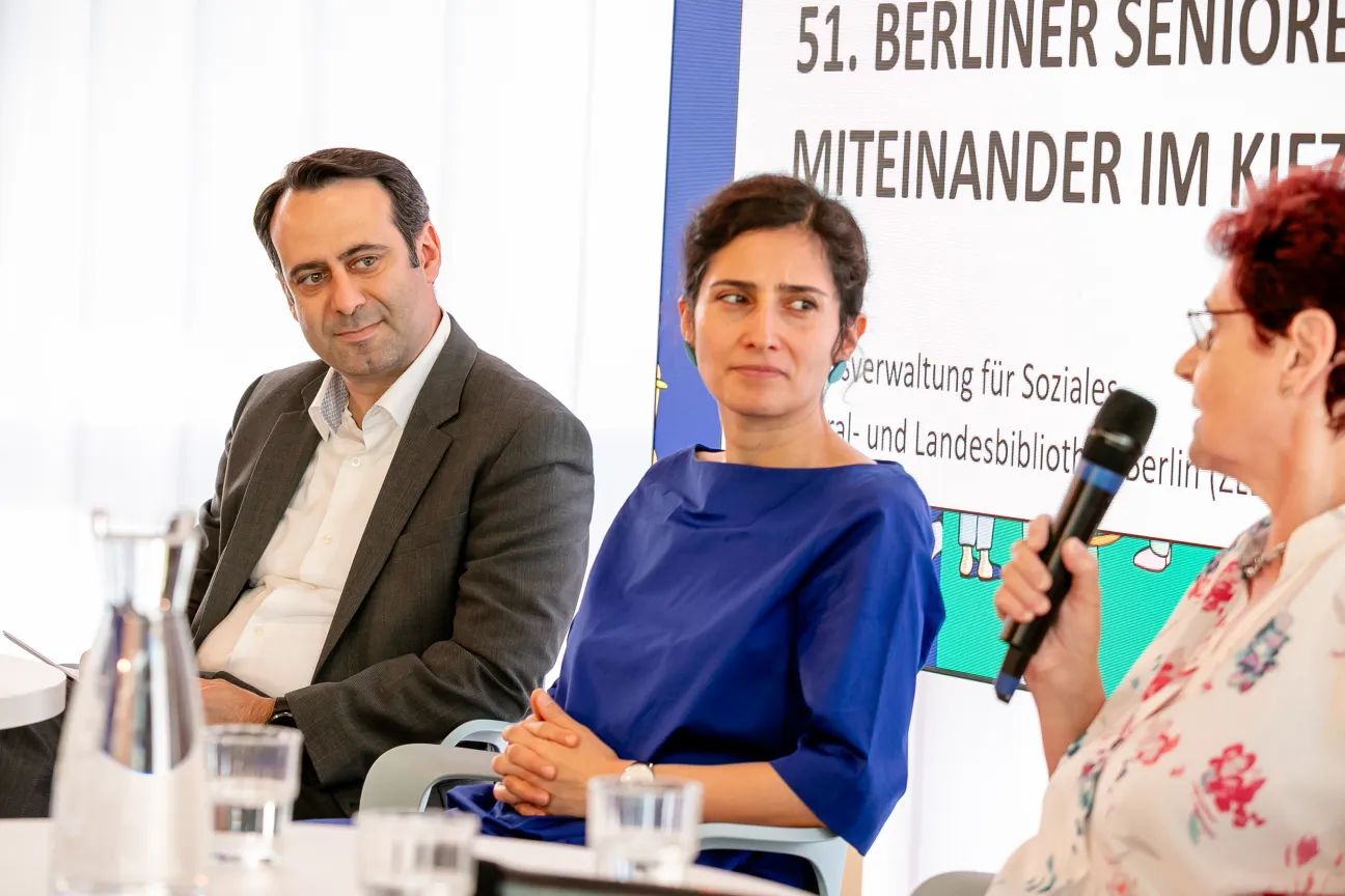 Drei Personen sitzen an einem Tisch. Im Hintergund ist zu lesen: 51. Berliner Seniorenwoche - Miteinander im Kiez. Eine Frau hält ein Mikrofon in der Hand, ein junge Frau und ein Mann sehen sie freundlich an. 