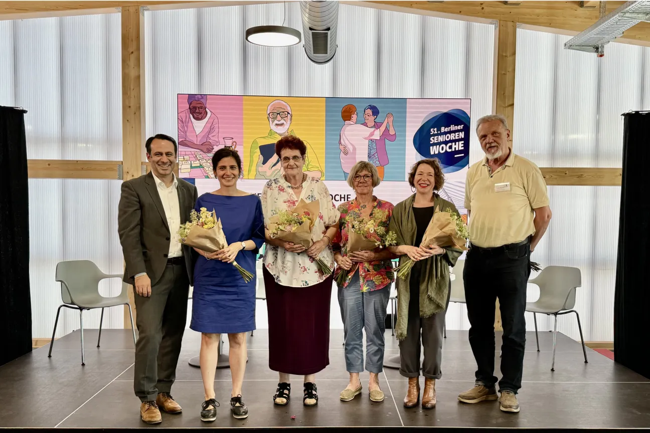 Blick auf eine Bühne mit lächelnden Personen: 4 Frauen mit Blumensträußen, 2 Männer. Dahinter bunte Grafiken zur 51. Berliner Seniorenwoche.